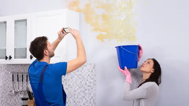 Homeowner photographing a water stain on a wall while another holds a bucket